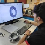 Ingénieur utilisant un logiciel CAO pour concevoir une pièce mécanique, avec un prototype physique posé à côté.